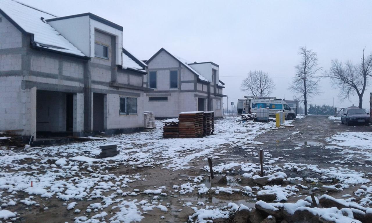 Surowy stan dwóch budynków w trakcie budowy, częściowo pokrytych śniegiem, z widocznym samochodem i przyczepą z reklamą na zewnątrz w pochmurny dzień.