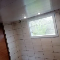 Wnętrze pomieszczenia z wykończonymi ścianami w jasnych, kwadratowych płytkach, białym oknem z widokiem na zieleń na zewnątrz, drewnianą podłogą i sufitem z paneli z wbudowanymi punktowymi lampkami.