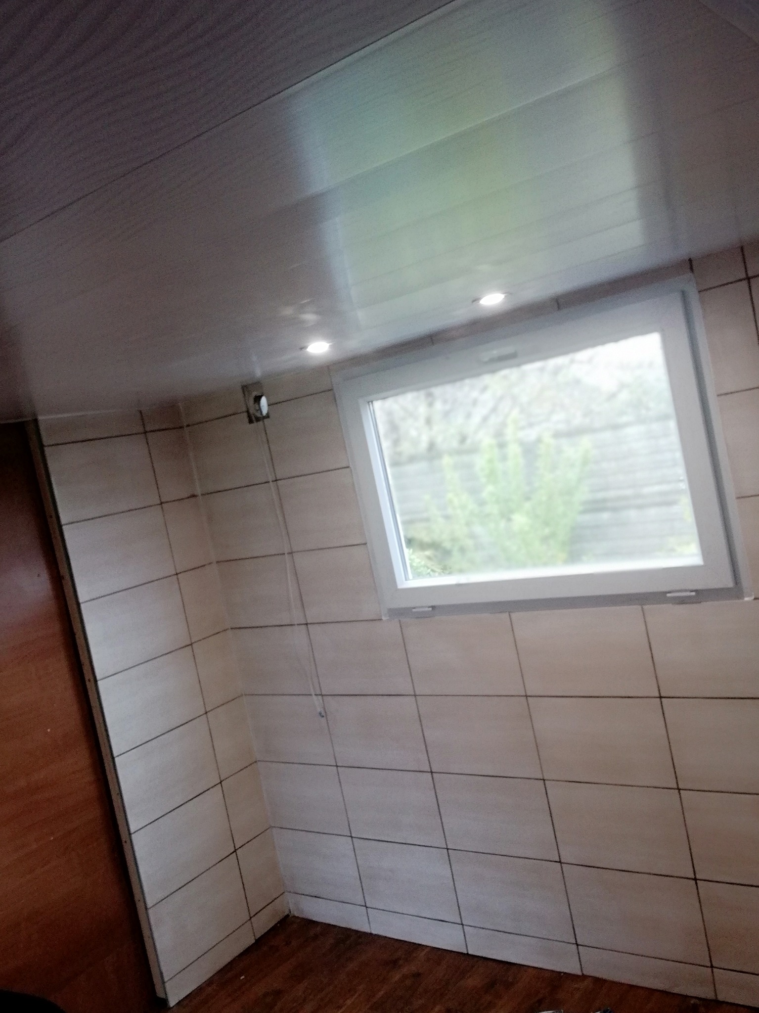 Wnętrze pomieszczenia z wykończonymi ścianami w jasnych, kwadratowych płytkach, białym oknem z widokiem na zieleń na zewnątrz, drewnianą podłogą i sufitem z paneli z wbudowanymi punktowymi lampkami.