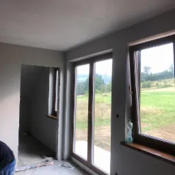 Wnętrze w trakcie prac wykończeniowych: świeżo nałożony tynk na ścianach, widoczne okna z drewnianą ramą i widokiem na zielone wzgórza, butelka wody na parapecie, częściowo widoczny pracownik...