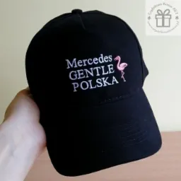 Czarna czapka z daszkiem, na której wyhaftowano biały napis 'Mercedes GENTLE POLSKA' oraz różowego flaminga, trzymana w dłoni.