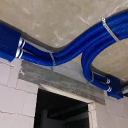 Niebieskie, karbowane rury wentylacyjne z białymi paskami, zamocowane do sufitu i ściany z bloków betonowych, widoczne metalowe uchwyty mocujące.