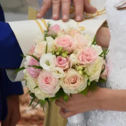 Panna młoda w białej sukni trzyma bukiet z różowych i białych róż, nad dłońmi narzeczonych ręka księdza.