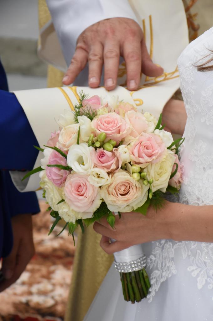 Panna młoda w białej sukni trzyma bukiet z różowych i białych róż, nad dłońmi narzeczonych ręka księdza.