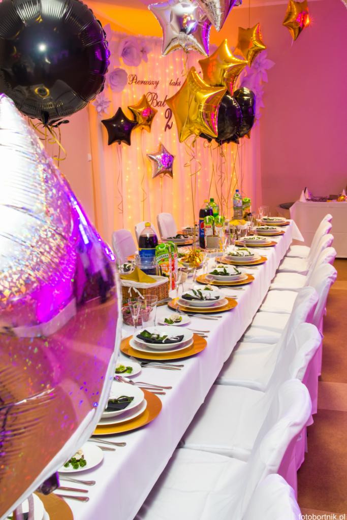 Uroczysty stół z białym obrusem, zastawą w kolorach złota i czerni, dekorowany balonami w kształcie gwiazd i kół, przygotowany na przyjęcie.