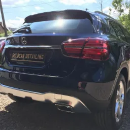 Błyszczący, granatowy Mercedes GLA 200 po detailingu, widok z tyłu, z tablicą rejestracyjną 'Goldcar Detailing', na żwirowym podłożu, w tle inne auta i budynki.