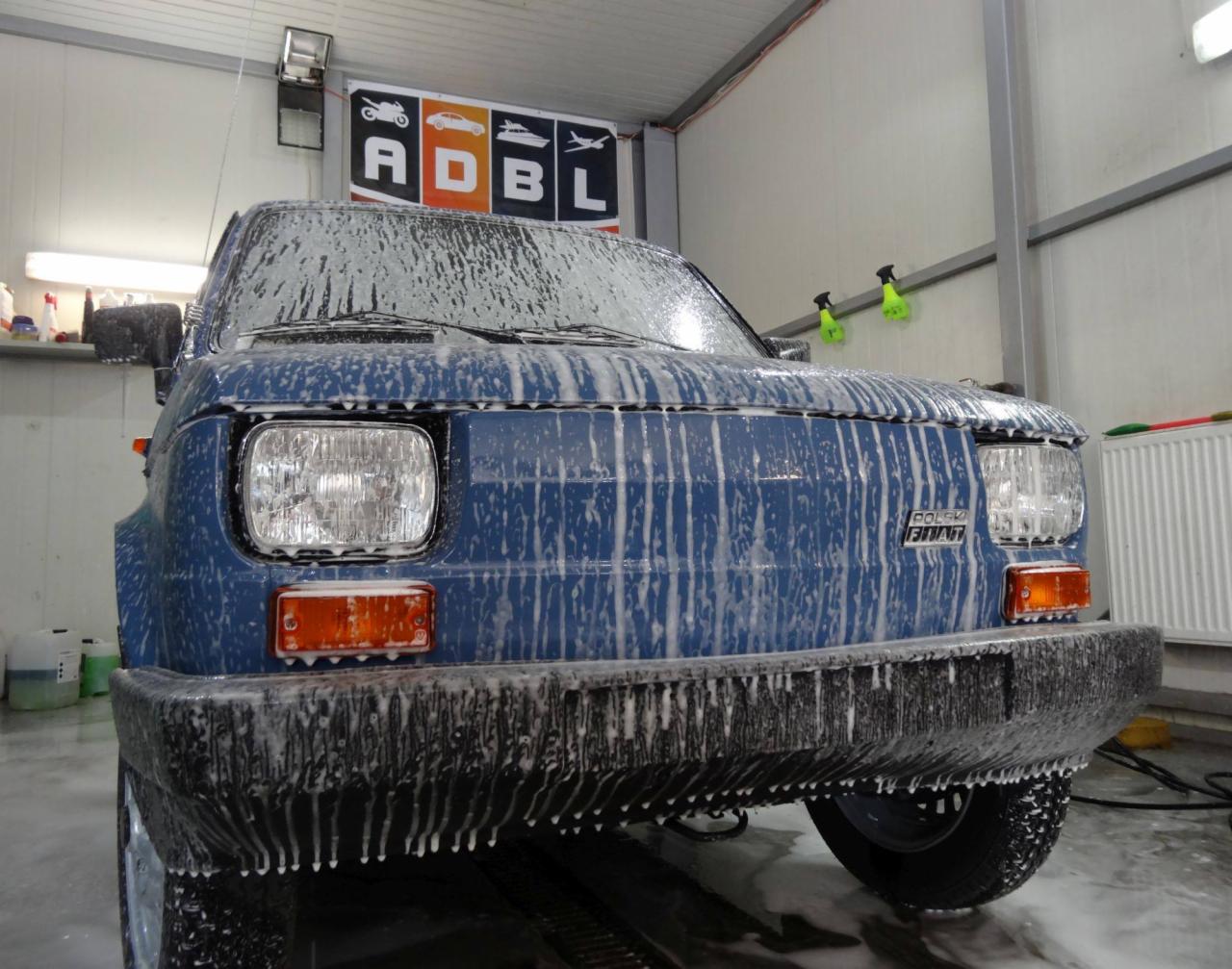 Niebieski Fiat 126p pokryty pianą podczas mycia w myjni samochodowej, widok z dołu, logo ADBL w tle.
