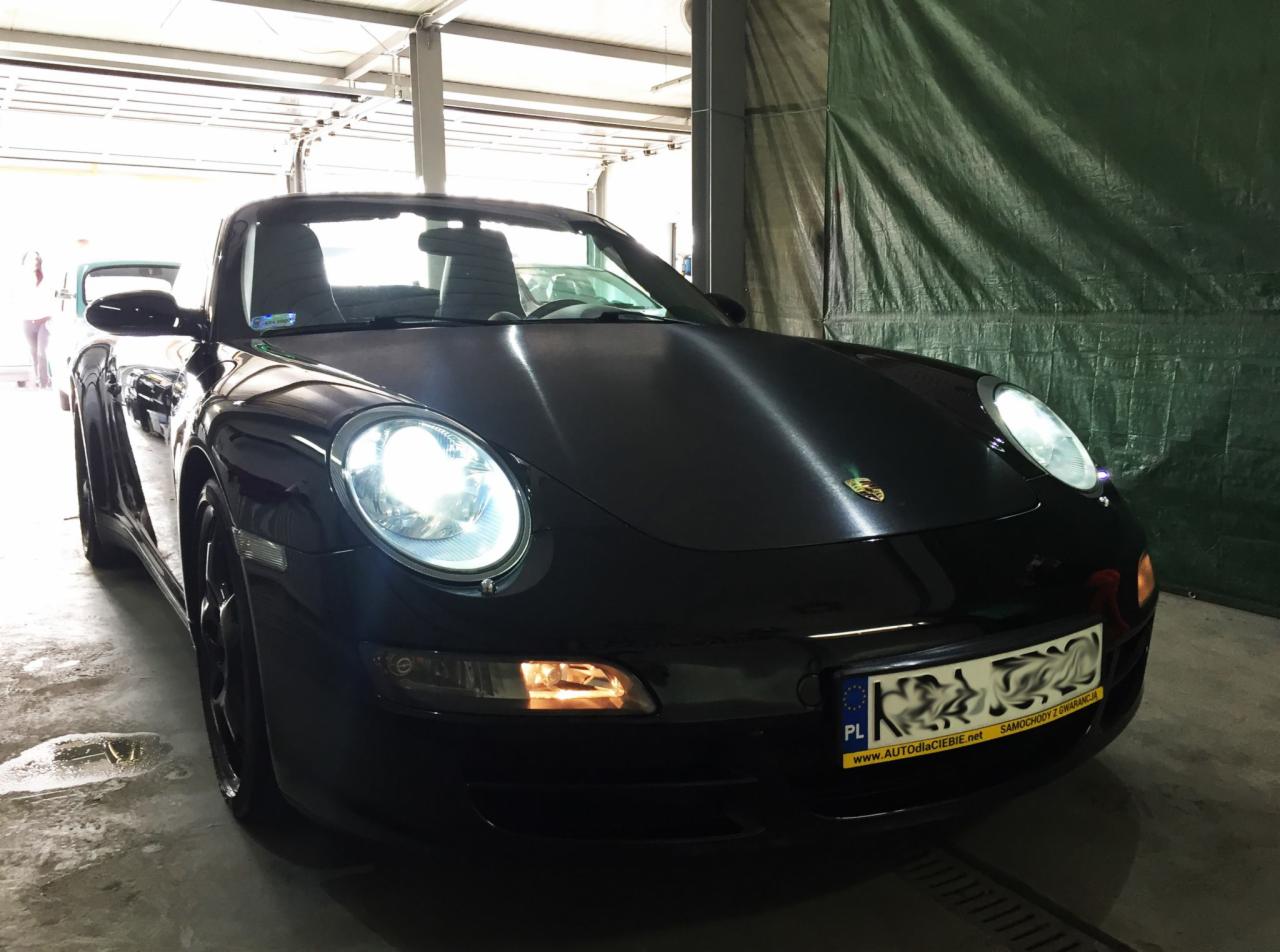 Czarny Porsche Cabriolet po umyciu, z włączonymi światłami, widoczny wewnątrz myjni samochodowej z zieloną plandeką w tle.