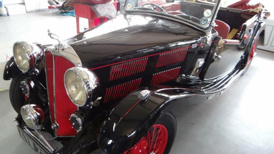 Czarny, zabytkowy kabriolet z czerwonymi akcentami, chromowanymi reflektorami i figurką orła na masce, zaparkowany w garażu.