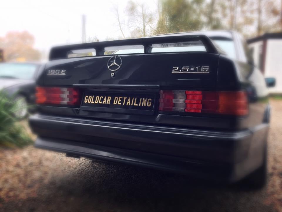 Czarny Mercedes-Benz 190E 2.5-16 po detailingu, widok z tyłu z tablicą rejestracyjną 'GOLDCAR DETAILING', w tle budynek i zieleń