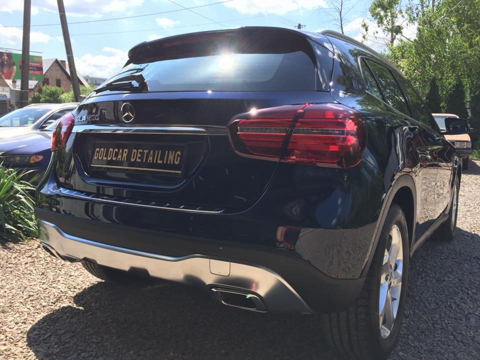 Błyszczący, granatowy Mercedes GLA 200 po detailingu, widok z tyłu, z tablicą rejestracyjną 'Goldcar Detailing', na żwirowym podłożu, w tle inne auta i budynki.