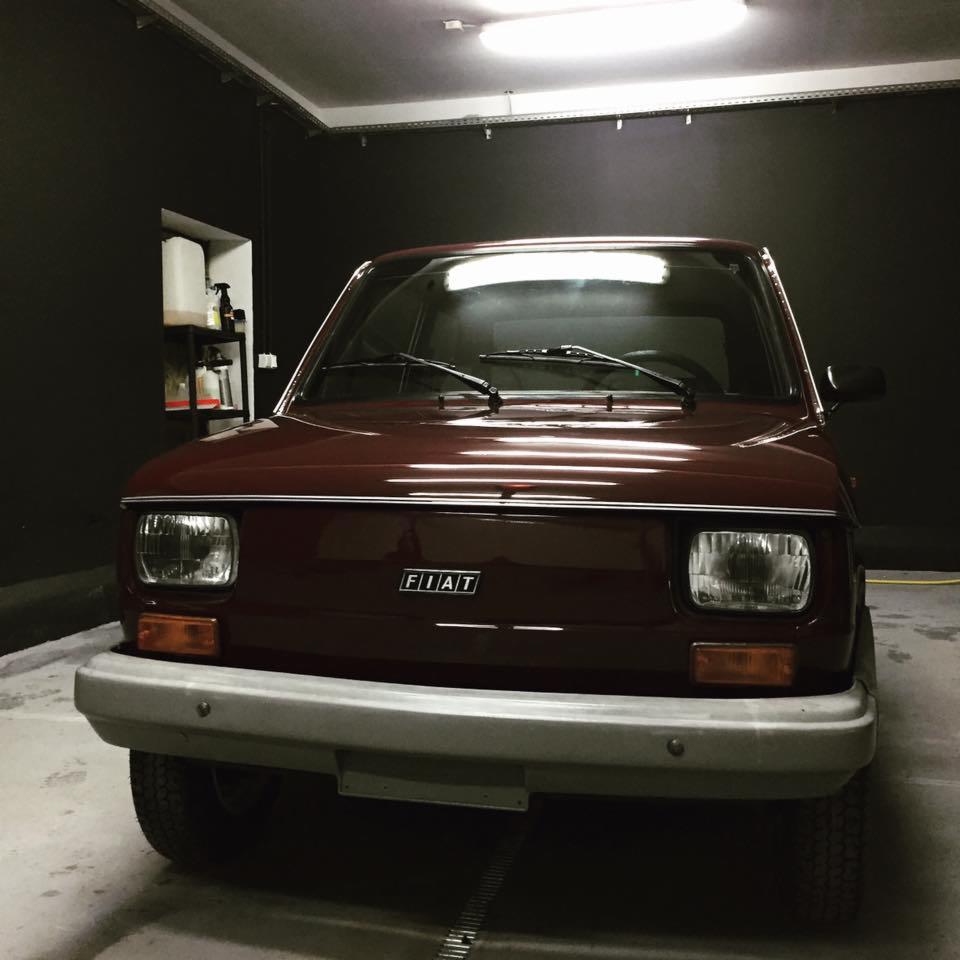 Przód bordowego Fiata 126p w garażu z czarnymi ścianami i widocznymi półkami z detergentami.