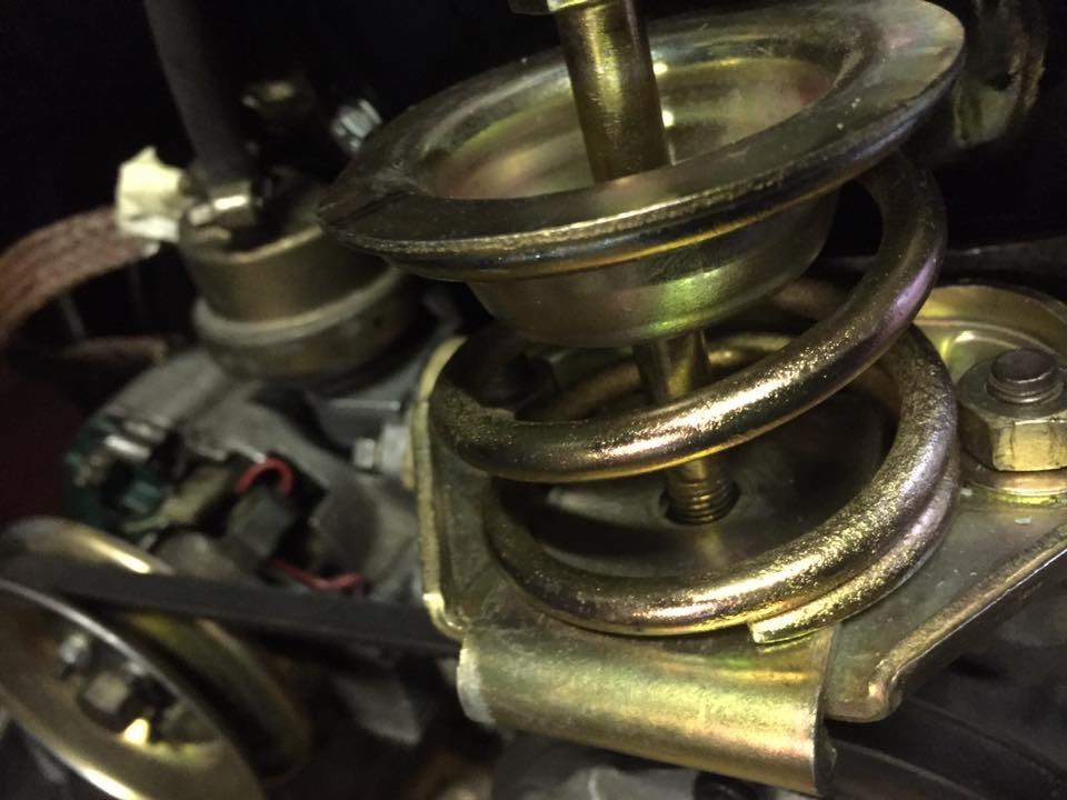 Szczegółowe ujęcie metalowej sprężyny zawieszenia samochodowego z widocznymi elementami mocującymi i paskiem napędowym w tle.