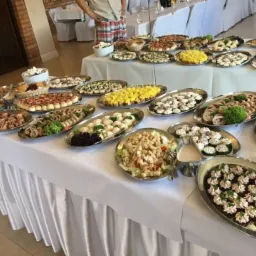 Stół szwedzki z różnorodnymi przekąskami na srebrnych tacach, w tle białe krzesła i osoba w kraciastej koszuli.