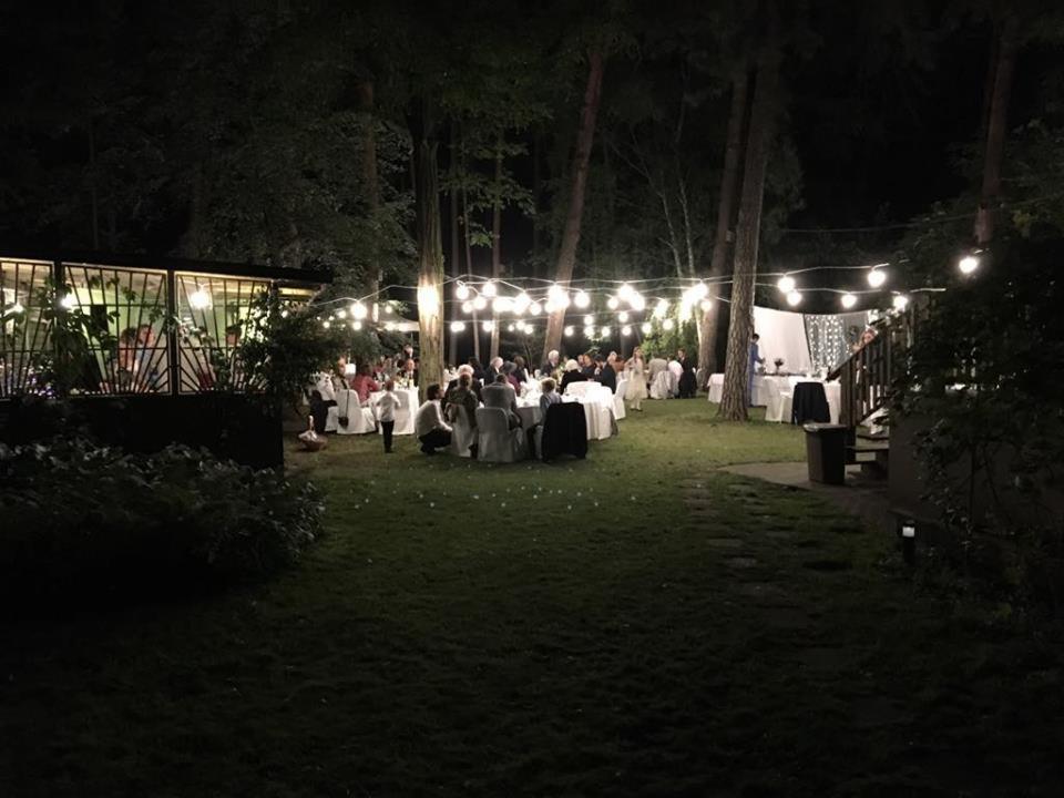 Uroczystość na zewnątrz, oświetlona girlandami żarówek, z gośćmi siedzącymi przy okrągłych stołach na trawniku, widoczny budynek z przeszkloną ścianą w tle.