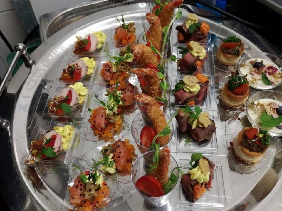 Srebrna taca z różnorodnymi przekąskami w przezroczystych naczyniach: krewetki w tempurze, roladki wołowe z kremem, tartaletki z farszem, łosoś z warzywami i mięso z truskawkami.