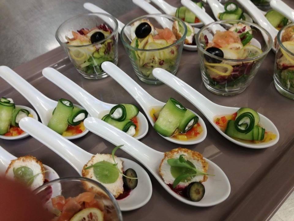 Eleganckie przekąski na białych łyżkach i w szklanych naczyniach, w tym roladki z ogórka z kremowym nadzieniem, smażony ser z ziołami i sałatki warzywne z owocami morza, ułożone na metalowej tacy.