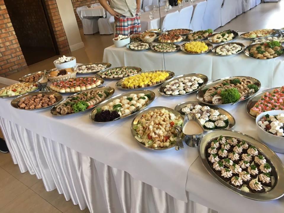 Stół szwedzki z różnorodnymi przekąskami na srebrnych tacach, w tle białe krzesła i osoba w kraciastej koszuli.