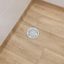 Jasna podłoga imitująca drewno w kabinie prysznicowej, z okrągłym odpływem w kolorze białym, widok z góry.