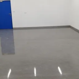 Gładka, szara posadzka w jasnym pomieszczeniu z białą ścianą i niebieskimi drzwiami, widoczne listwy przypodłogowe w kolorze szarym.