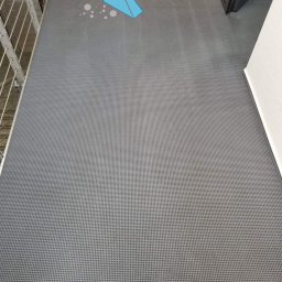 Santo Błysk - Szary dywan z drobną siatką, widoczny fragment balustrady i drzwi, na dywanie logo firmy sprzątającej z numerem telefonu.