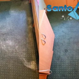 Zdjęcie dywanu z widoczną różnicą przed i po czyszczeniu, z logo firmy Santo Błysk i numerem telefonu widocznym w prawym górnym rogu, dywan zielony w małe białe kropki, obok szafka nocna.