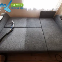 Szara rozkładana kanapa w trakcie czyszczenia, widoczna różnica w odcieniu po prawej stronie, dysza czyszcząca i wąż na kanapie, logo firmy Santo Błysk w górnym rogu.