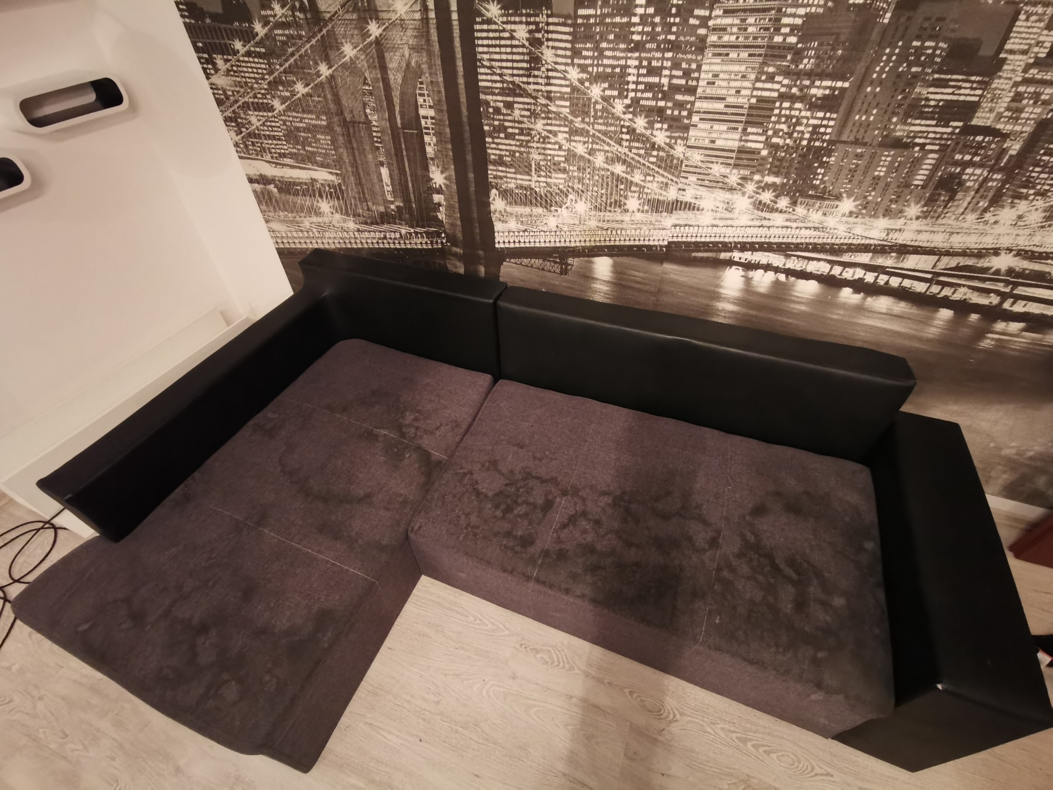 Szara, narożna kanapa z widocznymi plamami na tapicerce, na tle fototapety z motywem miasta nocą.