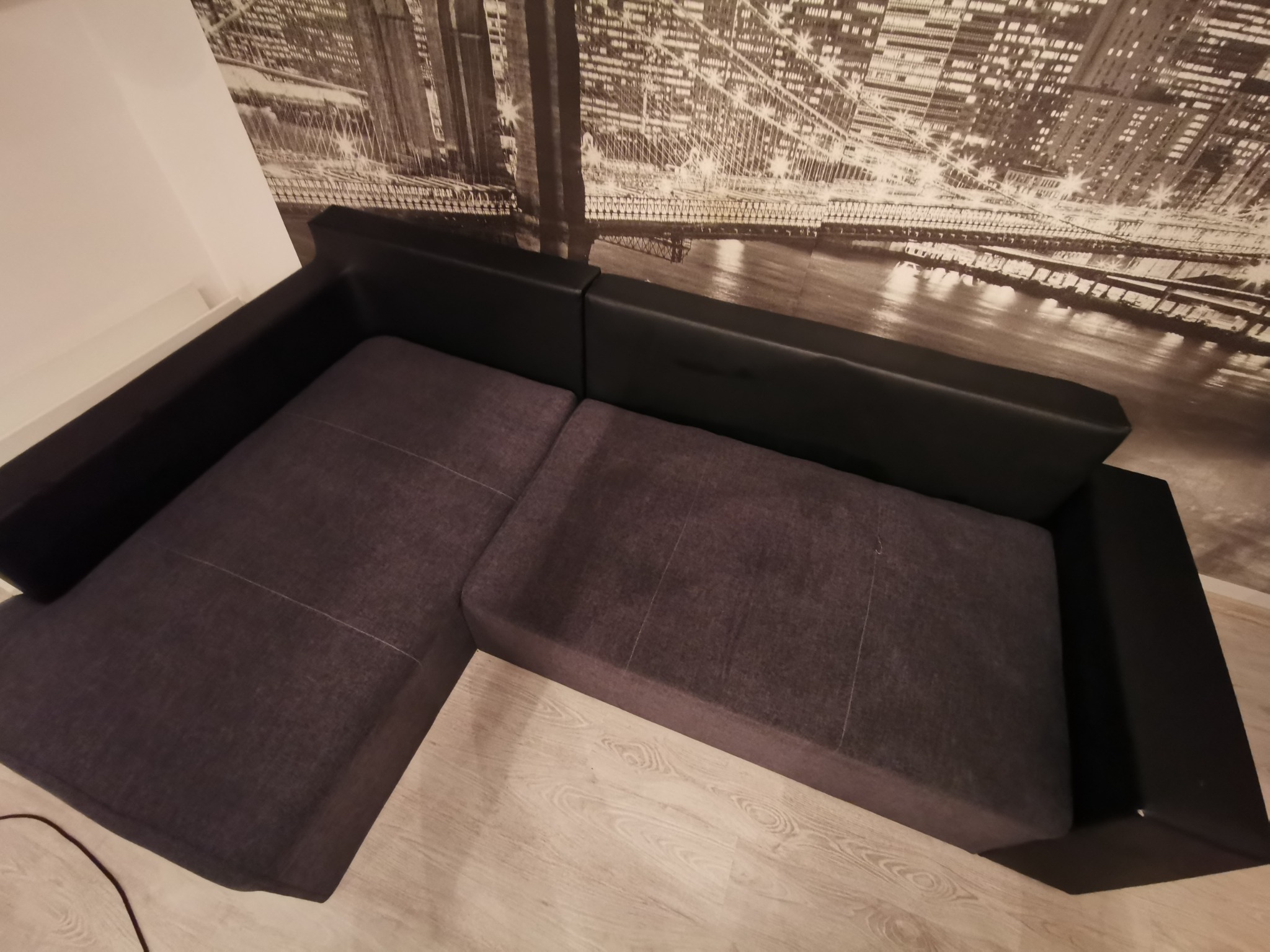 Szara, modułowa kanapa narożna z czarnymi bokami ustawiona na jasnej podłodze, na ścianie fototapeta z motywem miejskim.