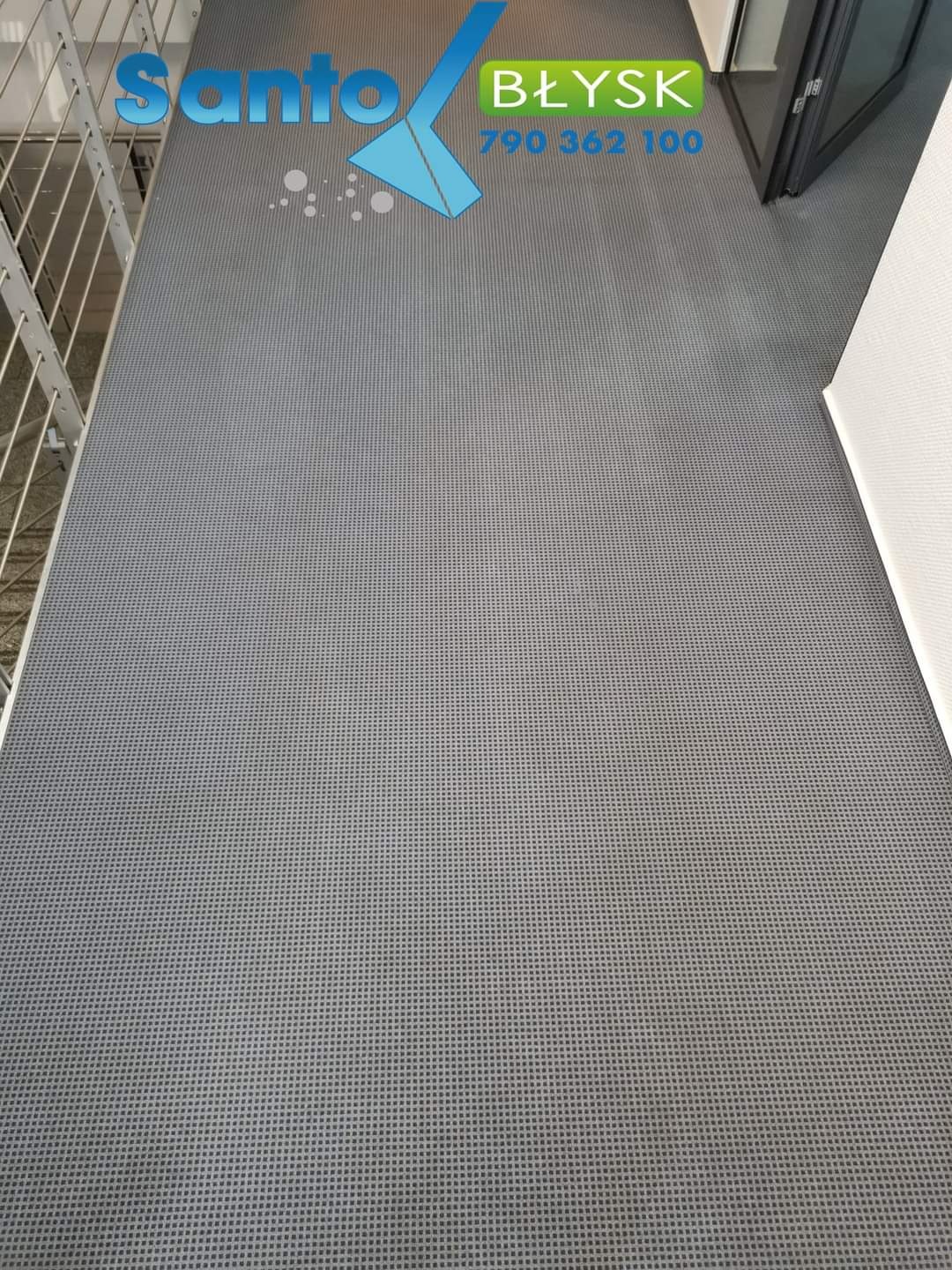 Szary dywan z drobną siatką, widoczny fragment balustrady i drzwi, na dywanie logo firmy sprzątającej z numerem telefonu.