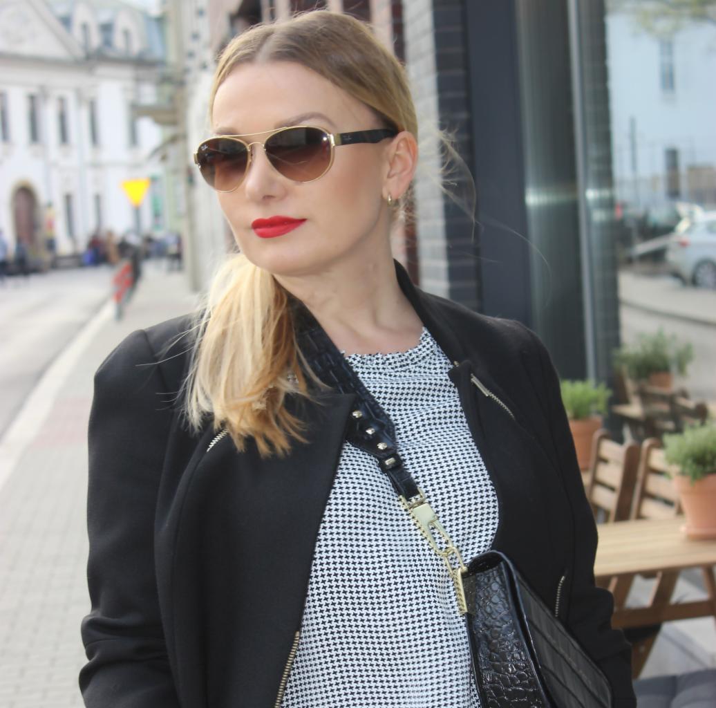 Blond kobieta w okularach przeciwsłonecznych z czerwoną szminką, ubrana w czarną marynarkę i wzorzystą bluzkę, pozująca na tle ulicy i kawiarnianych stolików.