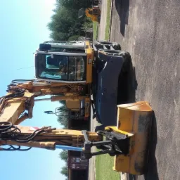 Żółta koparka kołowa na placu budowy w Piaskowcu, widoczna kabina operatora i ramię z łyżką, w tle inne maszyny budowlane.