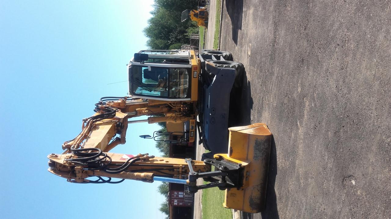 Żółta koparka kołowa na placu budowy w Piaskowcu, widoczna kabina operatora i ramię z łyżką, w tle inne maszyny budowlane.