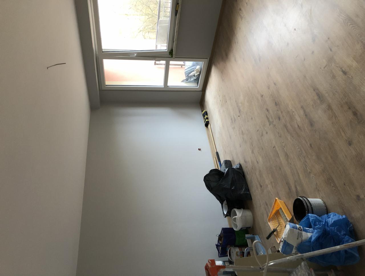 Pokój po malowaniu ścian na biało, widoczne narzędzia malarskie, wiadro z farbą i folia ochronna na podłodze z paneli.