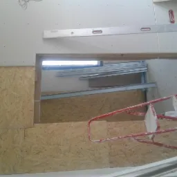 Montaż płyty karton gips na ścianie z widocznym otworem, częściowo zakrytej płytą OSB, z użyciem poziomicy i drabiny malarskiej.