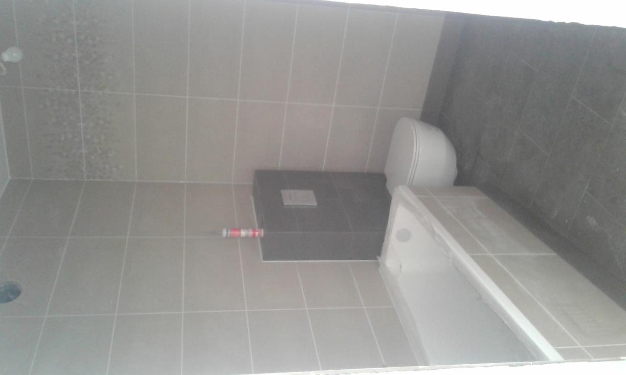 Wykończona łazienka z beżowymi płytkami ściennymi i podłogowymi, szarą zabudową WC, białą toaletą i brodzikiem z folią ochronną.