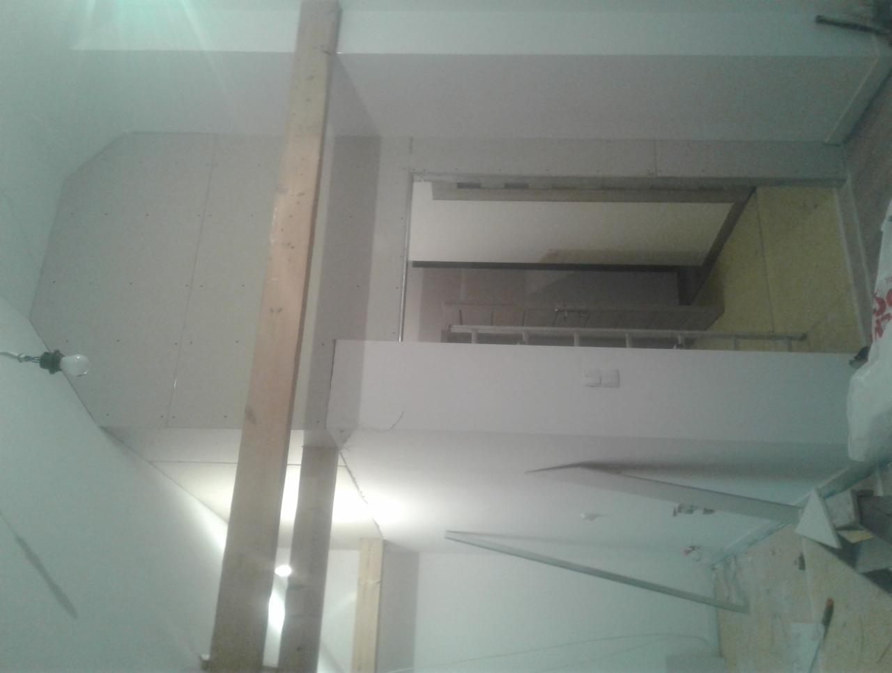 Montaż płyty kartonowo-gipsowej na ścianie, widoczna drewniana konstrukcja wspierająca, surowa żarówka zwisająca z sufitu, fragment drabiny w tle.