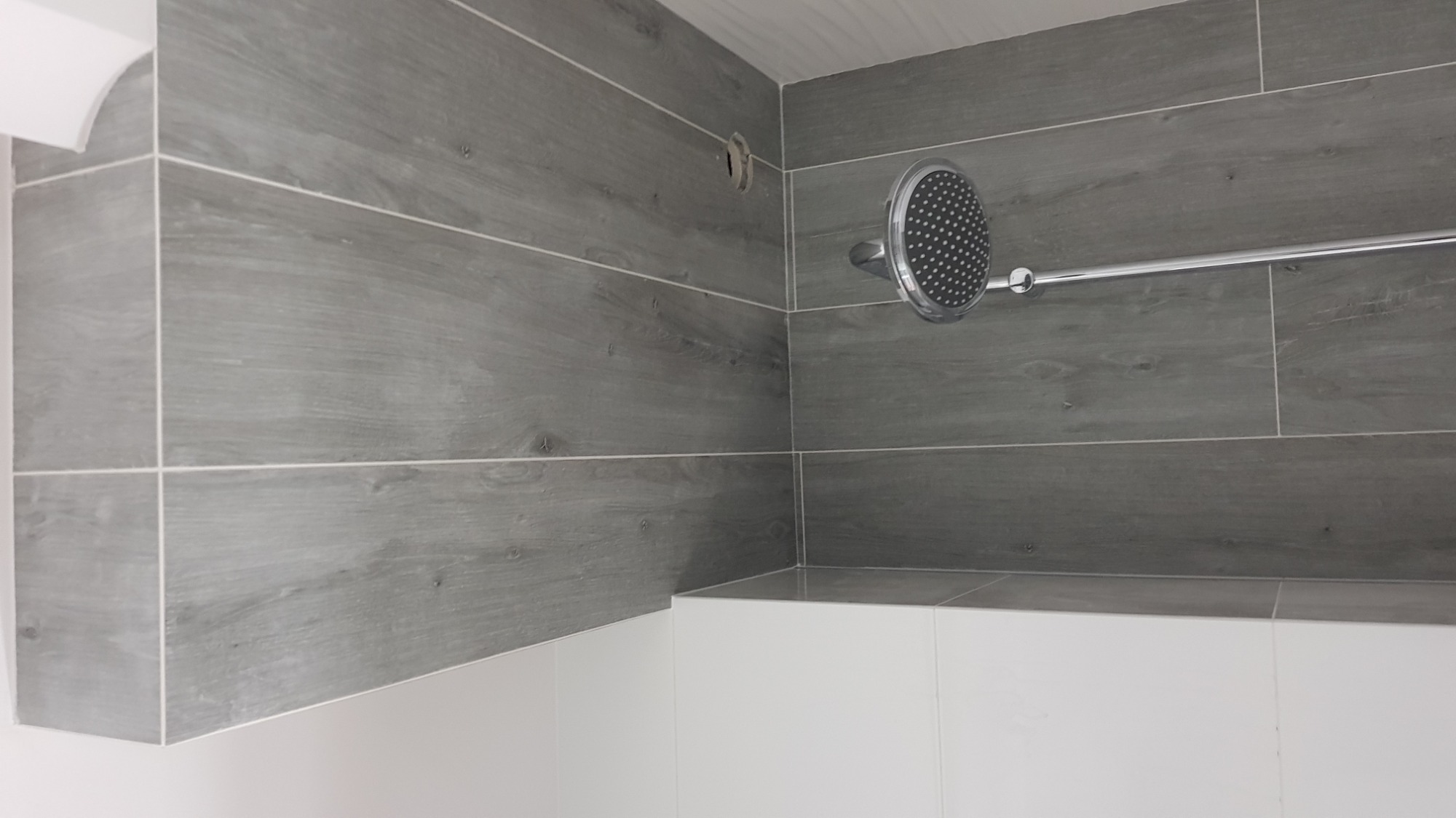 Nowoczesna łazienka z szarymi płytkami imitującymi drewno, chromowaną deszczownicą i białym obramowaniem wanny, widoczny otwór w ścianie przed montażem baterii.