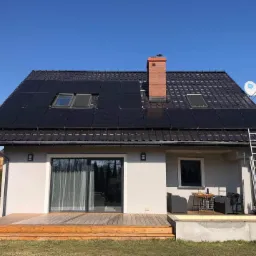 Dom jednorodzinny z panelami słonecznymi na dachu, ceglanym kominem i anteną satelitarną. Aluminiowa drabina oparta o rynnę. Widoczny taras z meblami ogrodowymi.