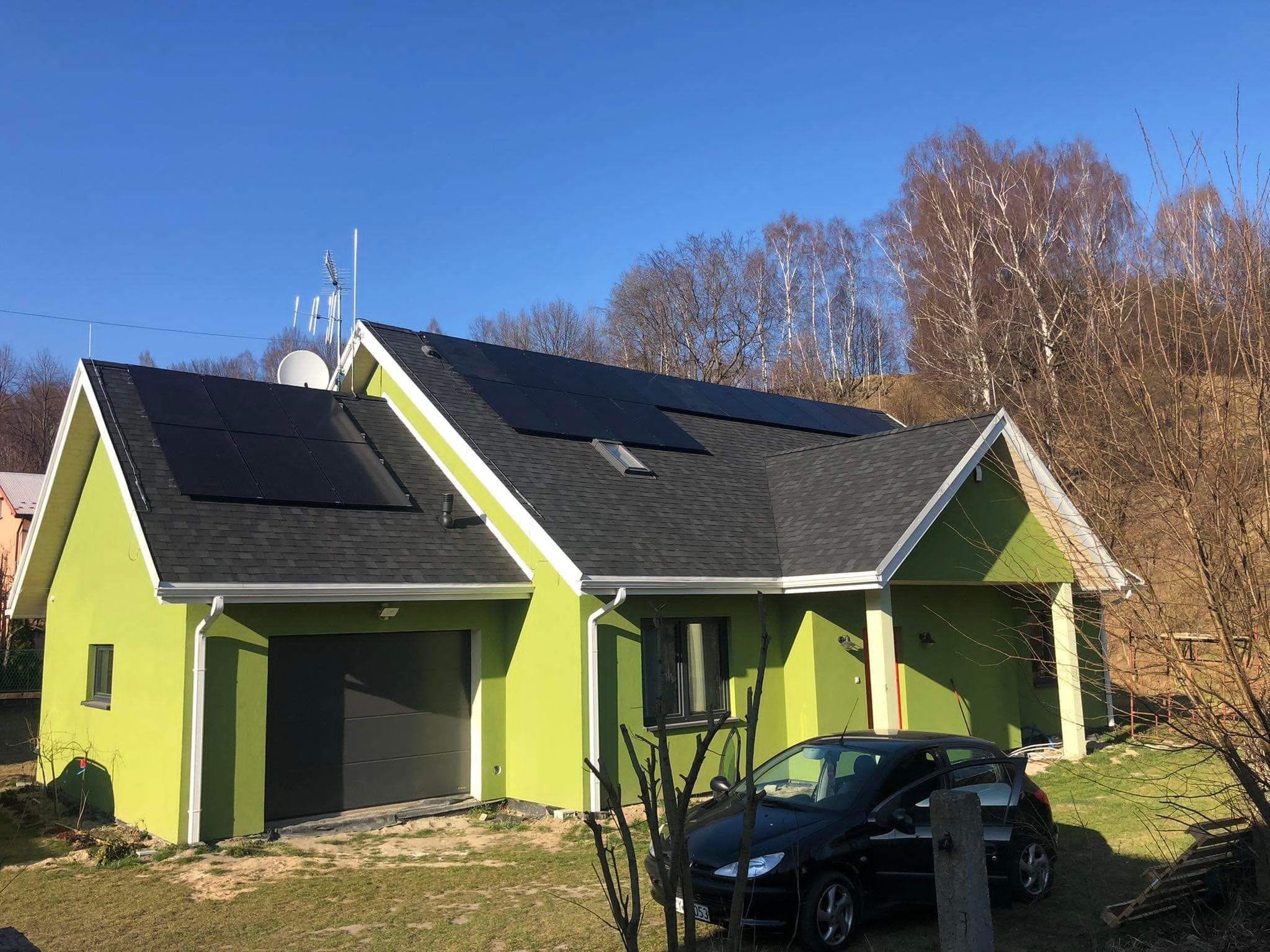 Dom jednorodzinny z zieloną elewacją i panelami fotowoltaicznymi na dachu pokrytym czarnym gontem bitumicznym, widoczny samochód osobowy.