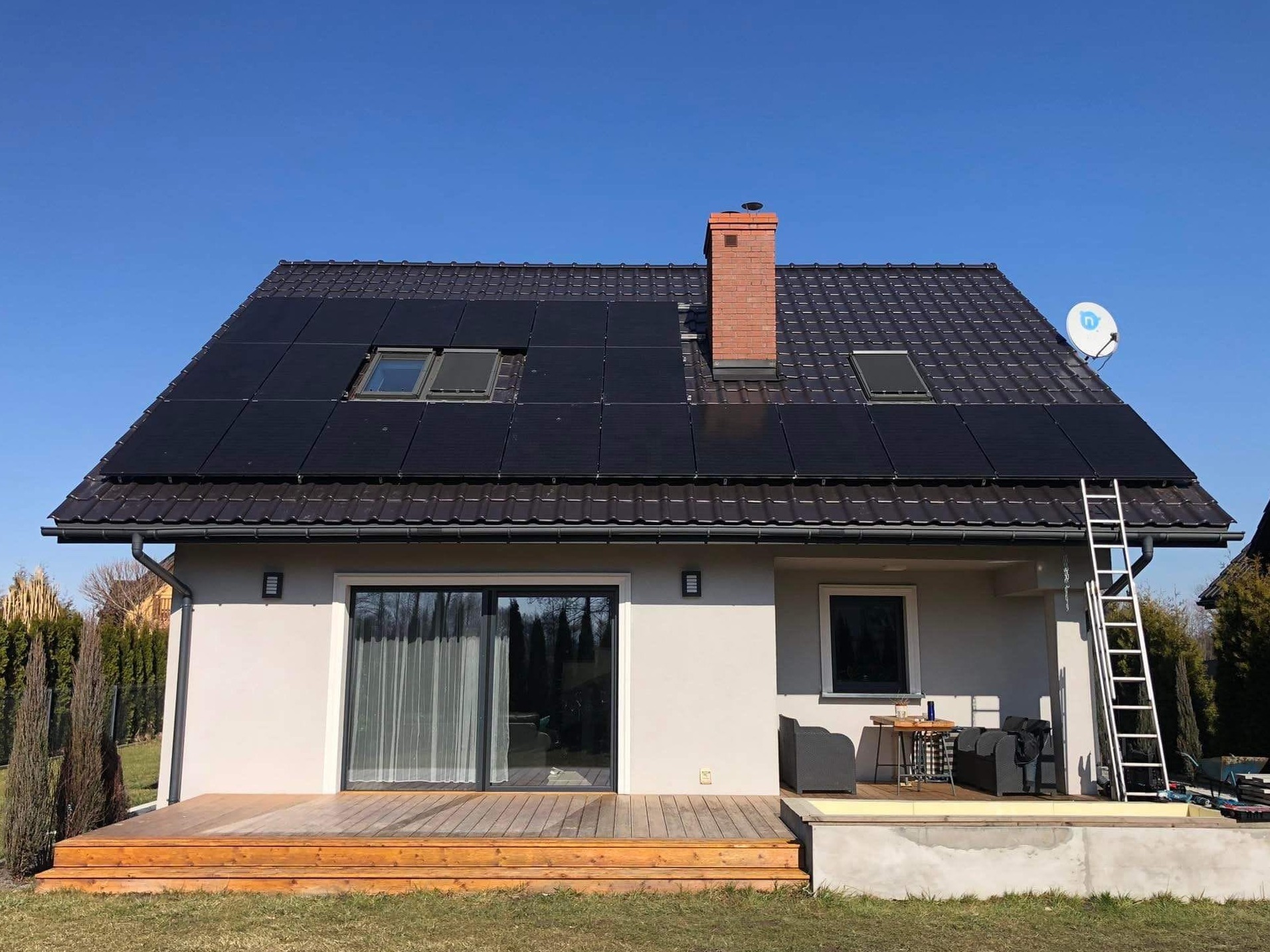Dom jednorodzinny z panelami słonecznymi na dachu, ceglanym kominem i anteną satelitarną. Aluminiowa drabina oparta o rynnę. Widoczny taras z meblami ogrodowymi.