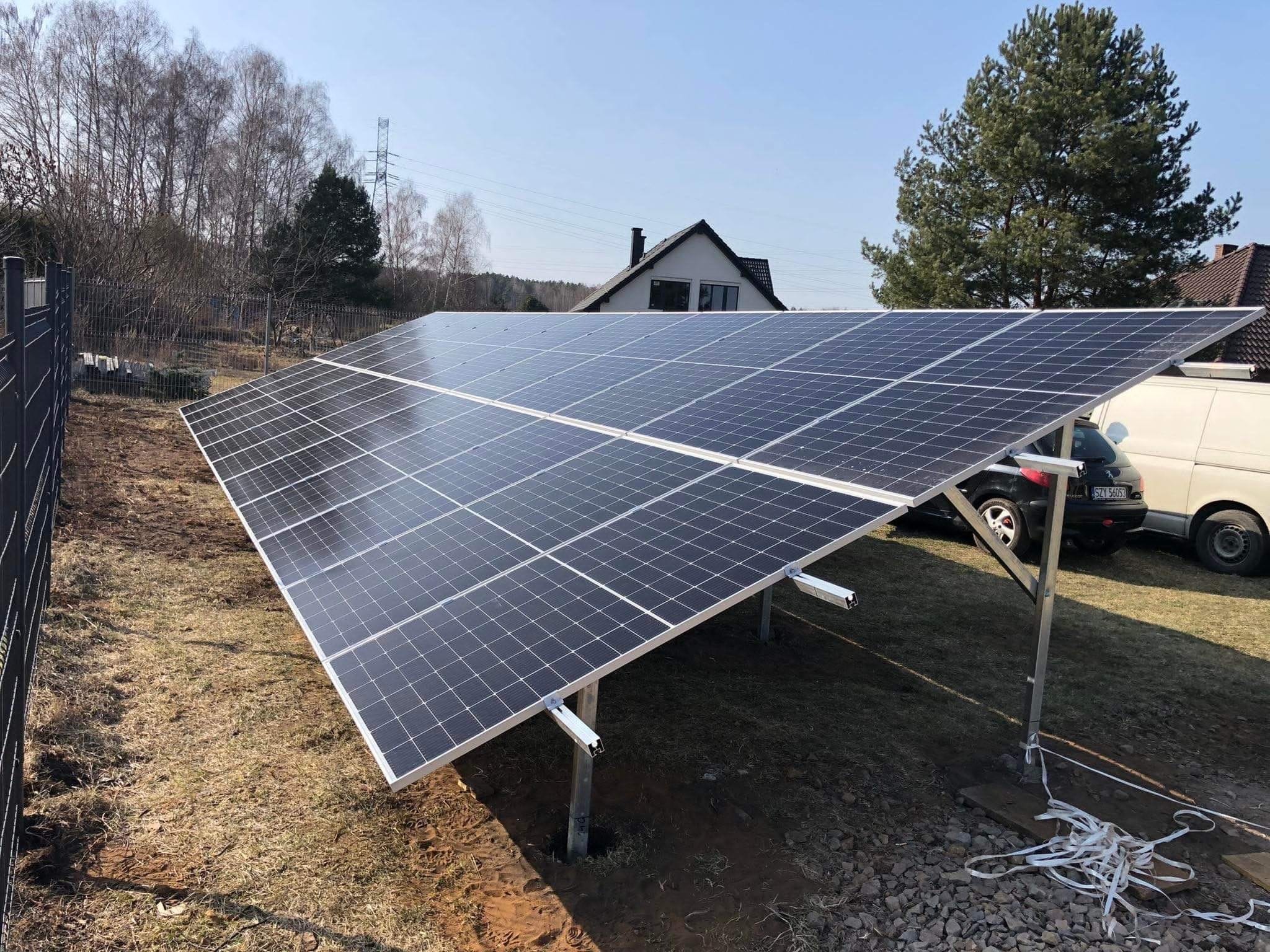 Duży, naziemny system paneli słonecznych ustawiony na metalowych wspornikach na trawiastym terenie, z widocznymi samochodami i domem w tle.