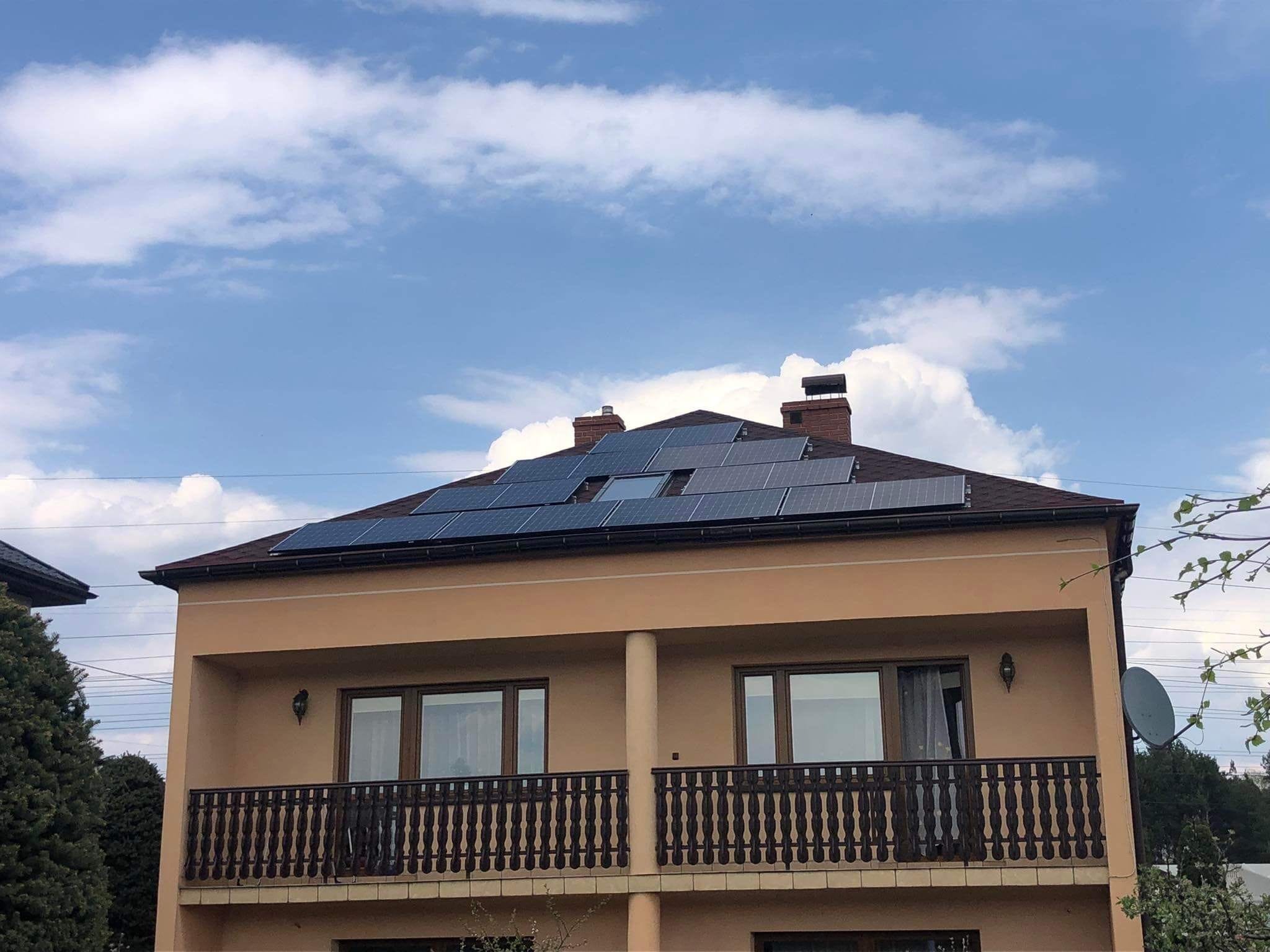 Dwupiętrowy dom z beżową elewacją i ciemnobrązowym dachem, na którym zamontowano panele słoneczne; widok z dołu na tle jasnego nieba z białymi chmurami.