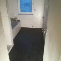 Wnętrze wykończonej łazienki z czarną posadzką i białymi płytkami na ścianach, widoczna wanna z baterią i okno z niebieską szybą, ujęcie z perspektywy wejścia.