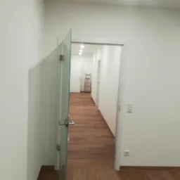 Wnętrze wyremontowanego mieszkania z drewnianą podłogą, widok na korytarz z otwartymi, szklanymi drzwiami i drabinką w tle, białe ściany i sufit z oświetleniem punktowym.