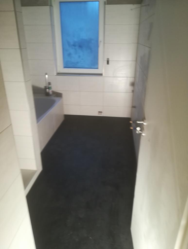 Wnętrze wykończonej łazienki z czarną posadzką i białymi płytkami na ścianach, widoczna wanna z baterią i okno z niebieską szybą, ujęcie z perspektywy wejścia.