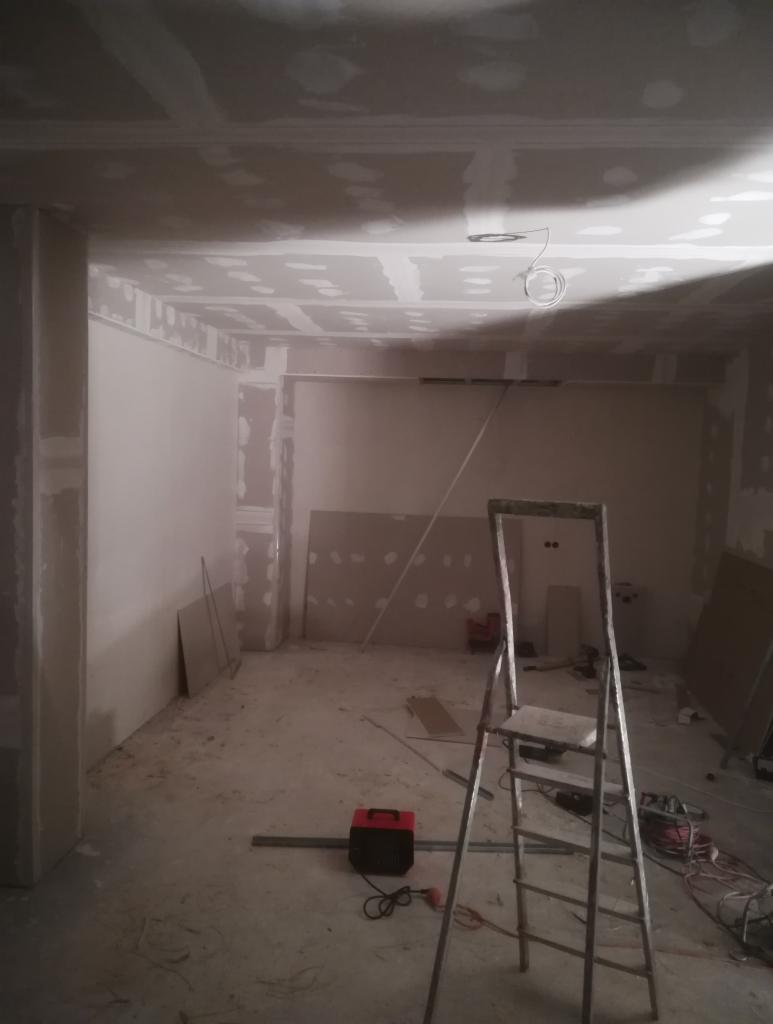 Wnętrze w trakcie wykańczania, ściany i sufit pokryte płytami, widoczna drabina, przewody elektryczne zwisające z sufitu, oraz narzędzia na podłodze.