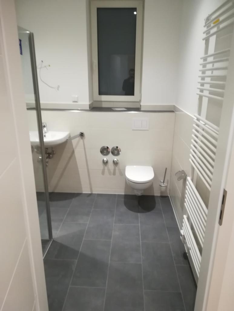 Nowoczesna łazienka z szarymi płytkami podłogowymi, białymi ścianami i sanitariatami. W oknie odbija się sylwetka osoby.