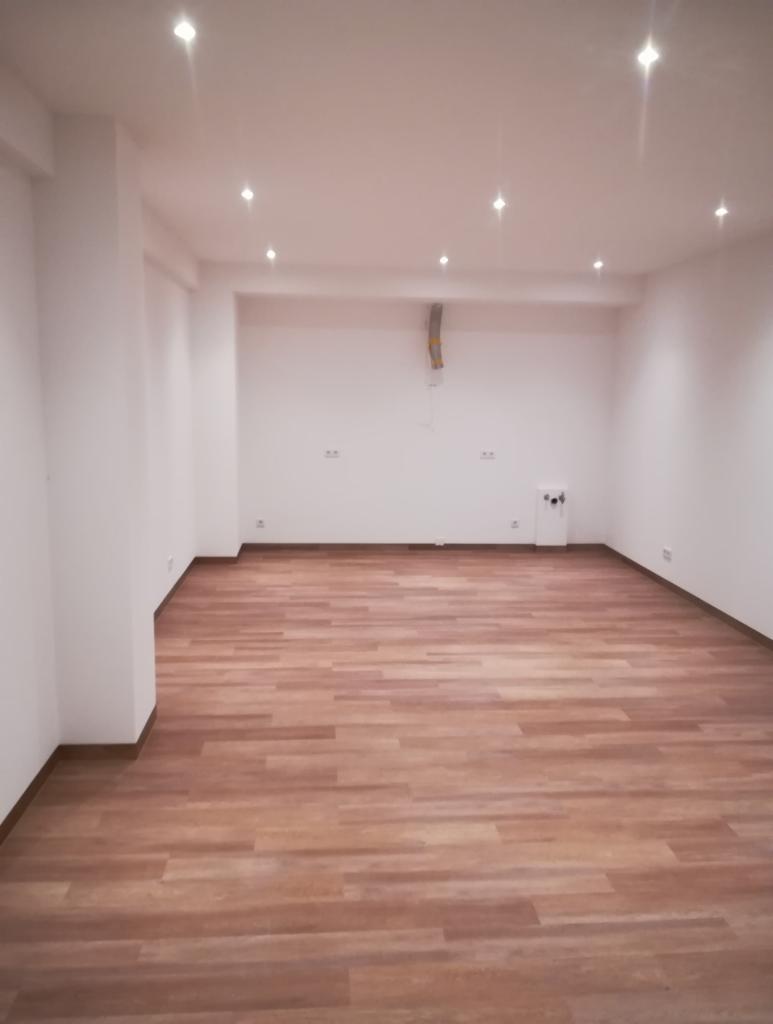 Puste pomieszczenie po remoncie z nową podłogą z paneli w odcieniach brązu, białe ściany i sufit z punktowym oświetleniem halogenowym. Widoczne gniazdka elektryczne. Na suficie wystający fragment...