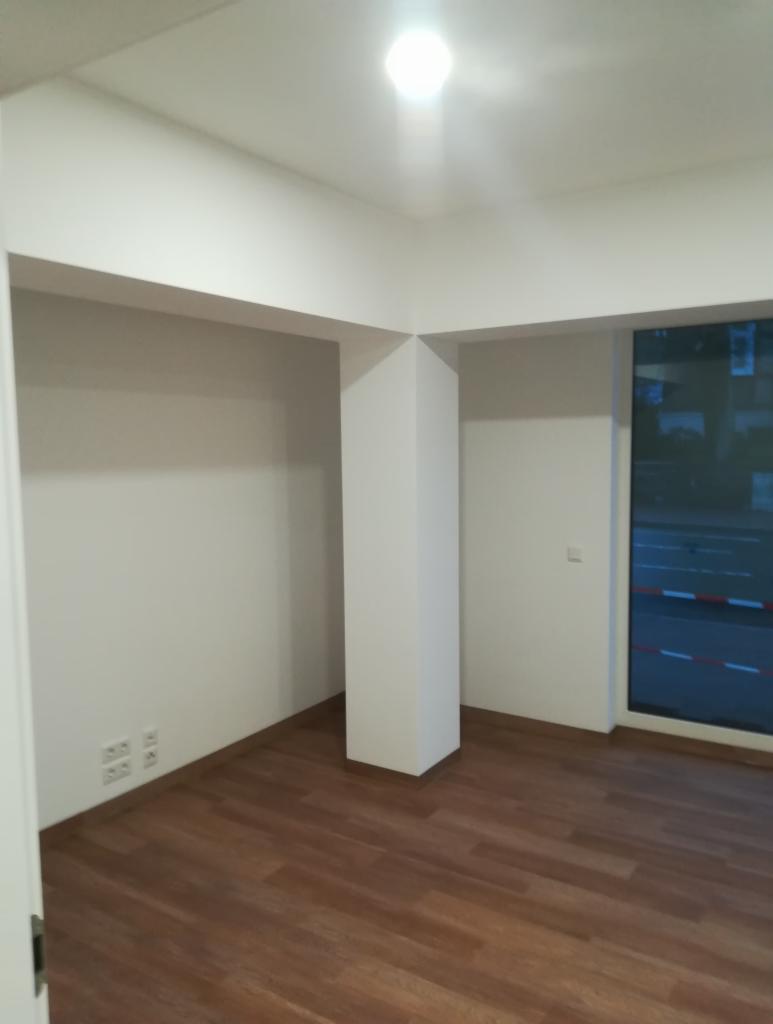 Pusty pokój z brązowymi panelami podłogowymi, białe ściany i sufit, widoczna kolumna konstrukcyjna i okno z widokiem na ulicę.